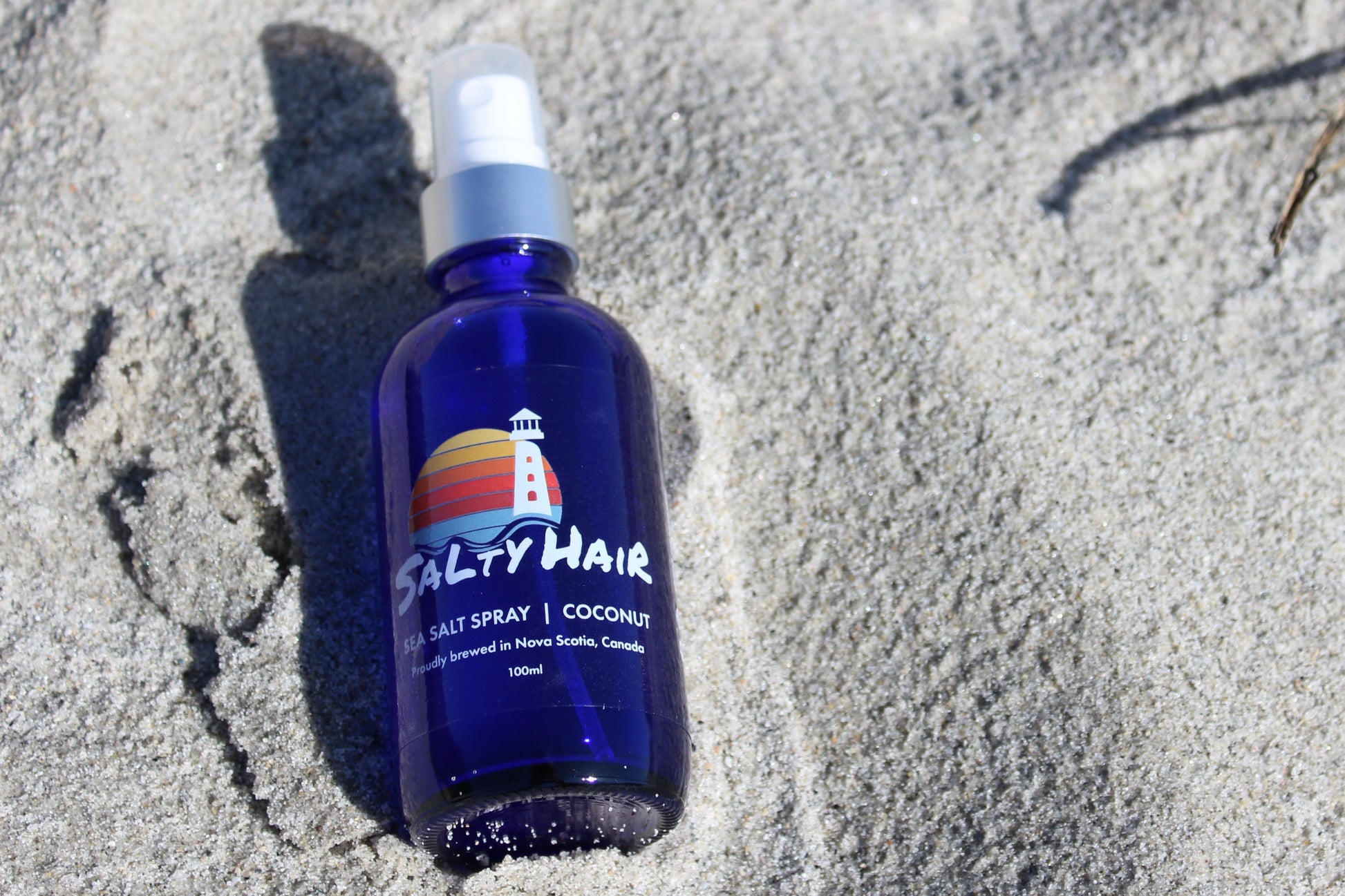 A blue glass bottle of coconut-scented Salty Hair sea salt spray, sitting in sand at the beach.