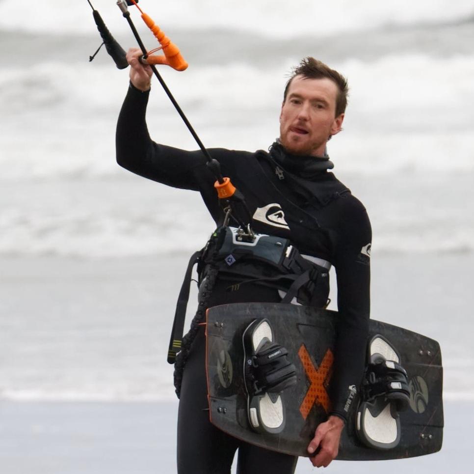 Sustainable hair company Salty Hair founder Mike Petrosoniak walking on a Nova Scotia beach wearing a wetsuit while carrying his kiteboarding gear.