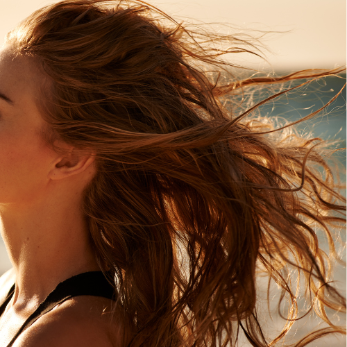 Woman with windswept long hair standing on the beach, highlighting styling tips for long hair with Salty Hair Sea Salt Spray.